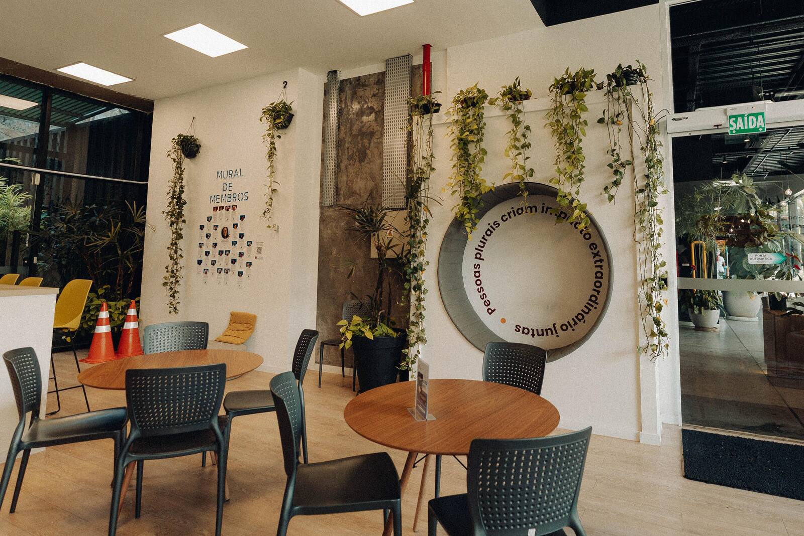 Área de convivência do Impact Hub Floripa com mesas redondas, plantas e o Mural de Membros